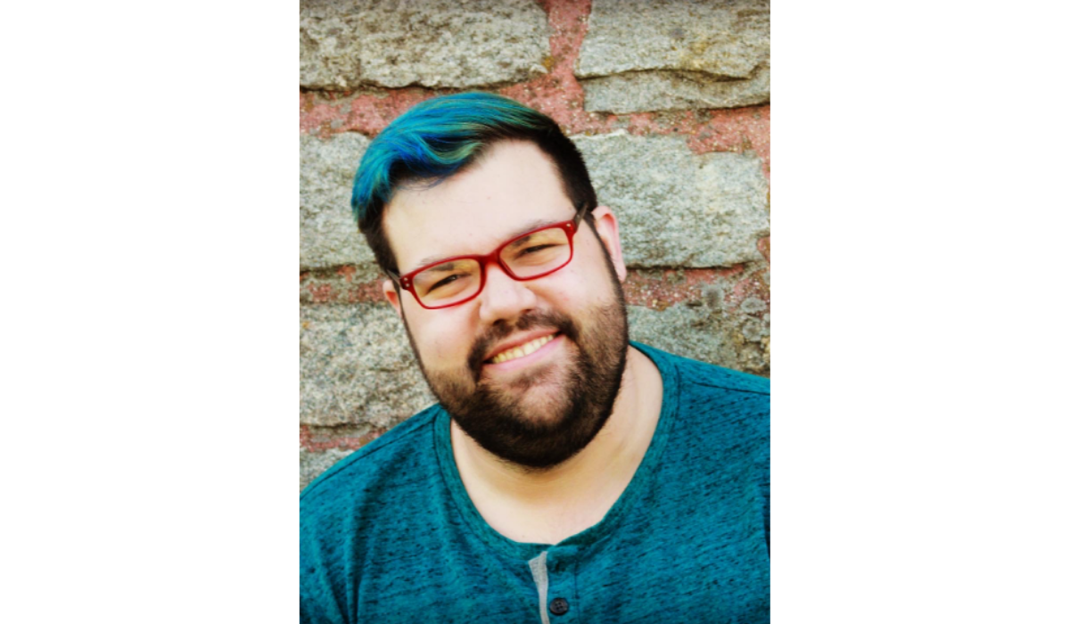 A close-up photograph of a smiling, bearded person wearing red-framed glasses and a teal shirt. They have dark hair with streaks of bright blue and green color visible on the top and right side of their head. The background is a textured wall made of large, weathered stones.