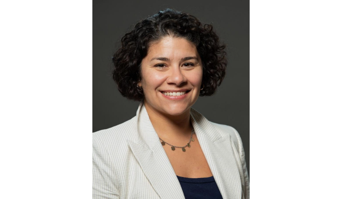 A headshot of a smiling woman with short, dark, curly hair. She is wearing a light-colored blazer, possibly white or cream with a subtle vertical stripe, over a dark shirt. She is also wearing a delicate necklace with small, round charms. The background is a solid, dark gray.
