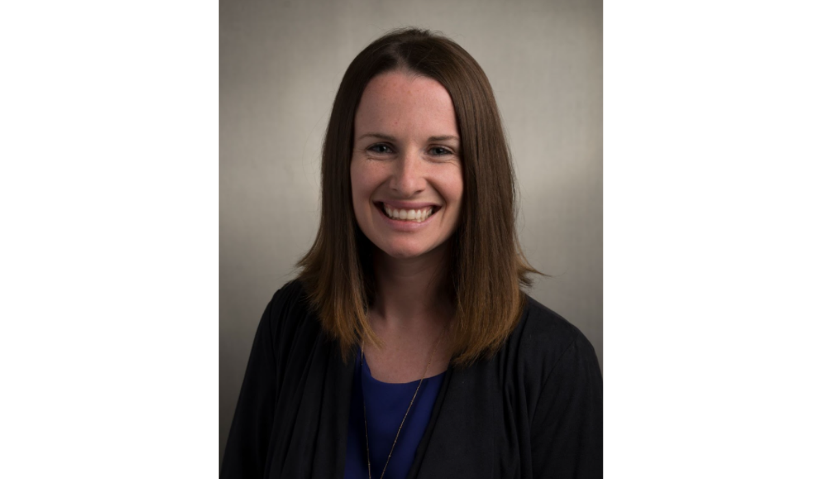 A headshot of a smiling woman with straight, shoulder-length brown hair parted in the middle. She is wearing a black cardigan or open sweater over a bright blue top, and a long, thin gold pendant necklace. The background is a soft, light gray or beige color with subtle shading.